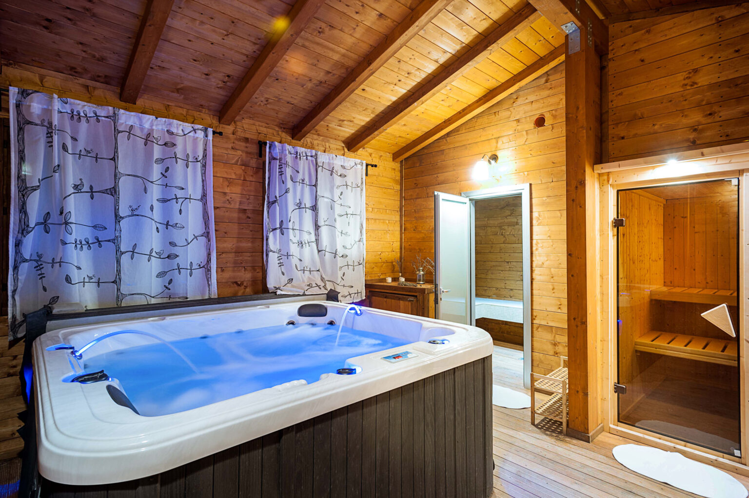 Cozy wooden spa room featuring a luxurious hot tub with blue lighting, surrounded by decorative curtains and wooden paneling, alongside a sauna and a relaxing atmosphere. | Only on SopranoVillas