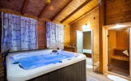 Cozy wooden spa room featuring a luxurious hot tub with blue lighting, surrounded by decorative curtains and wooden paneling, alongside a sauna and a relaxing atmosphere.