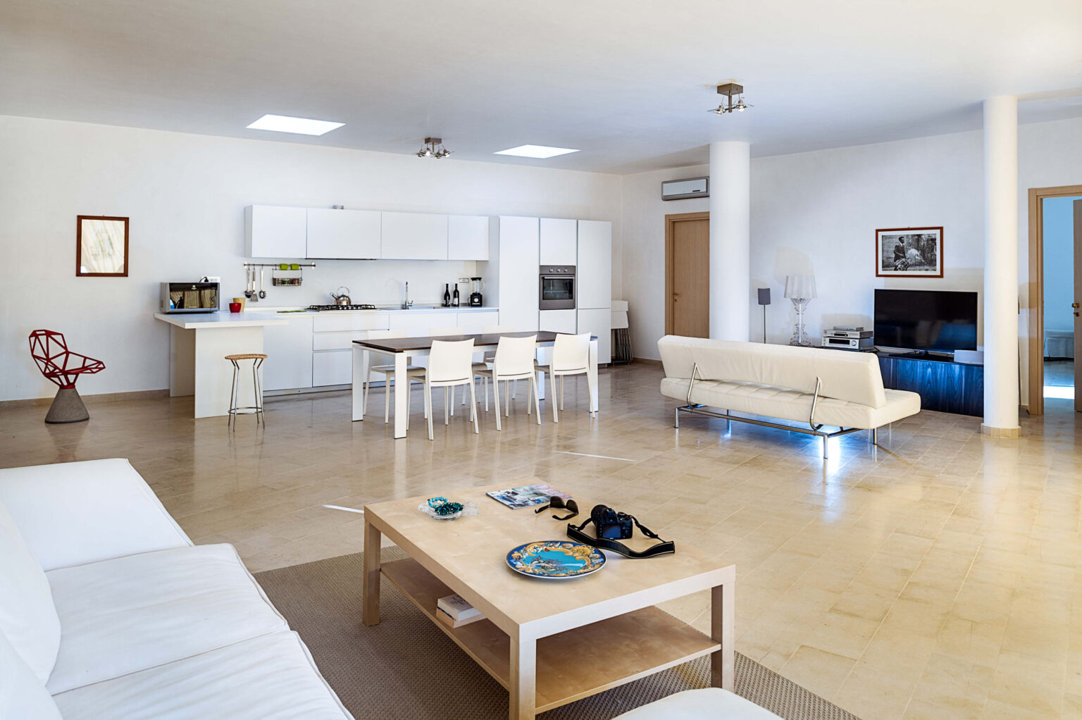 Spacious modern living room with a minimalist design, featuring a comfortable white sofa, a wooden coffee table, and a dining area with white chairs. The open kitchen is equipped with sleek cabinetry, and large windows allow natural light to fill the space. | Only on SopranoVillas