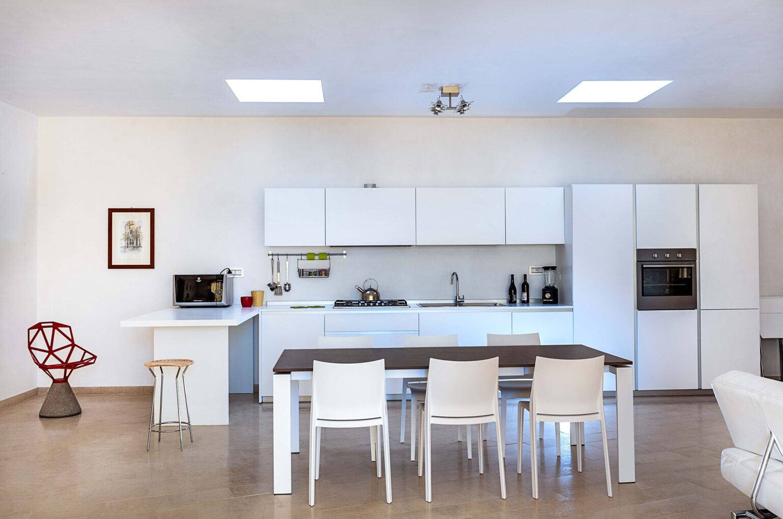 Modern kitchen interior featuring a minimalist design with white cabinetry, a sleek dining table, and stylish seating. The space is well-lit with natural light from skylights, showcasing appliances like a microwave, gas stove, and built-in oven. Decorative elements include a framed artwork and a unique red chair, adding a pop of color to the neutral palette. | Only on SopranoVillas