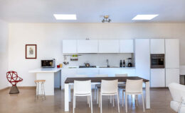Modern kitchen interior featuring a minimalist design with white cabinetry, a sleek dining table, and stylish seating. The space is well-lit with natural light from skylights, showcasing appliances like a microwave, gas stove, and built-in oven. Decorative elements include a framed artwork and a unique red chair, adding a pop of color to the neutral palette.