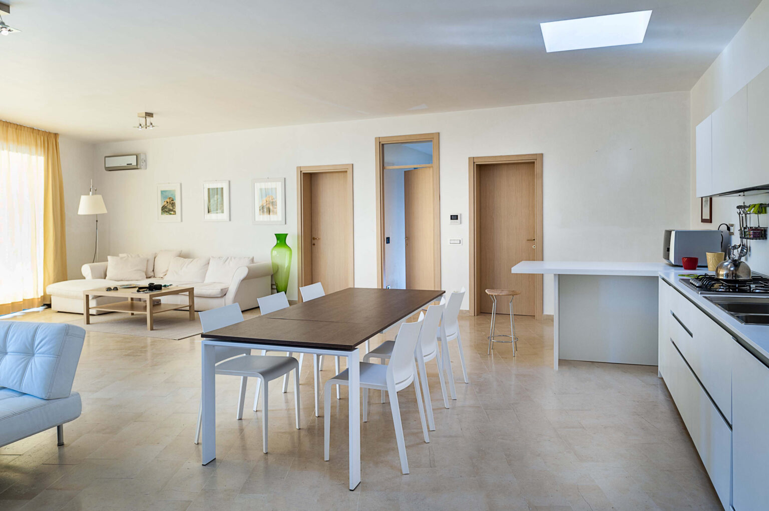 Modern open-concept living space featuring a dining table with white chairs, a cozy sofa, and a minimalist kitchen with sleek cabinetry and appliances. Natural light floods the room through large windows, creating a bright and inviting atmosphere. | Only on SopranoVillas
