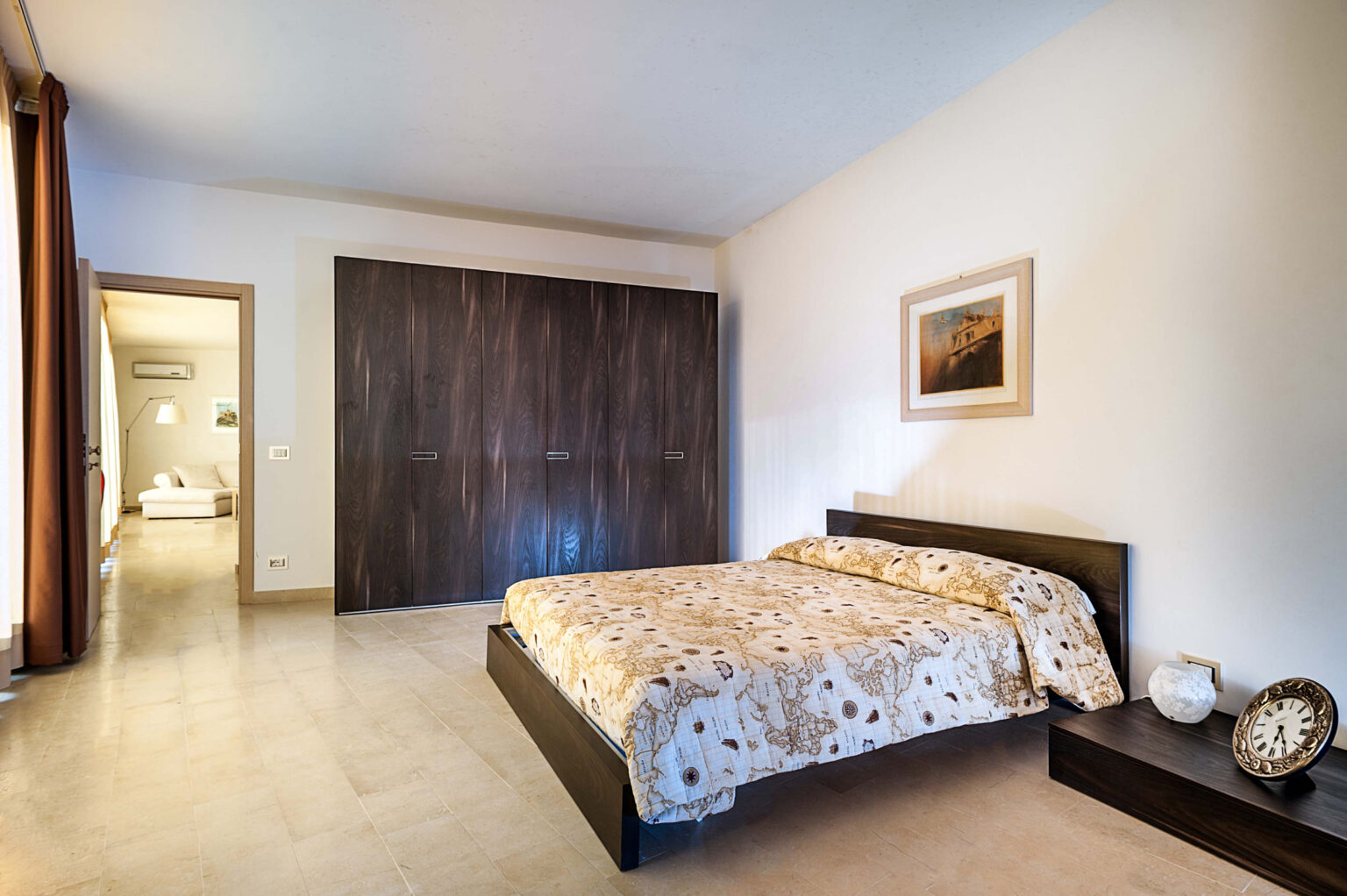Modern bedroom featuring a queen-sized bed with a patterned duvet, dark wooden furniture, and a minimalist design. The room includes a built-in wardrobe, a decorative wall clock, and a painting, with natural light streaming through the window, creating a cozy atmosphere. | Only on SopranoVillas