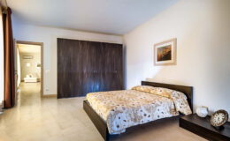 Modern bedroom featuring a queen-sized bed with a patterned duvet, dark wooden furniture, and a minimalist design. The room includes a built-in wardrobe, a decorative wall clock, and a painting, with natural light streaming through the window, creating a cozy atmosphere.