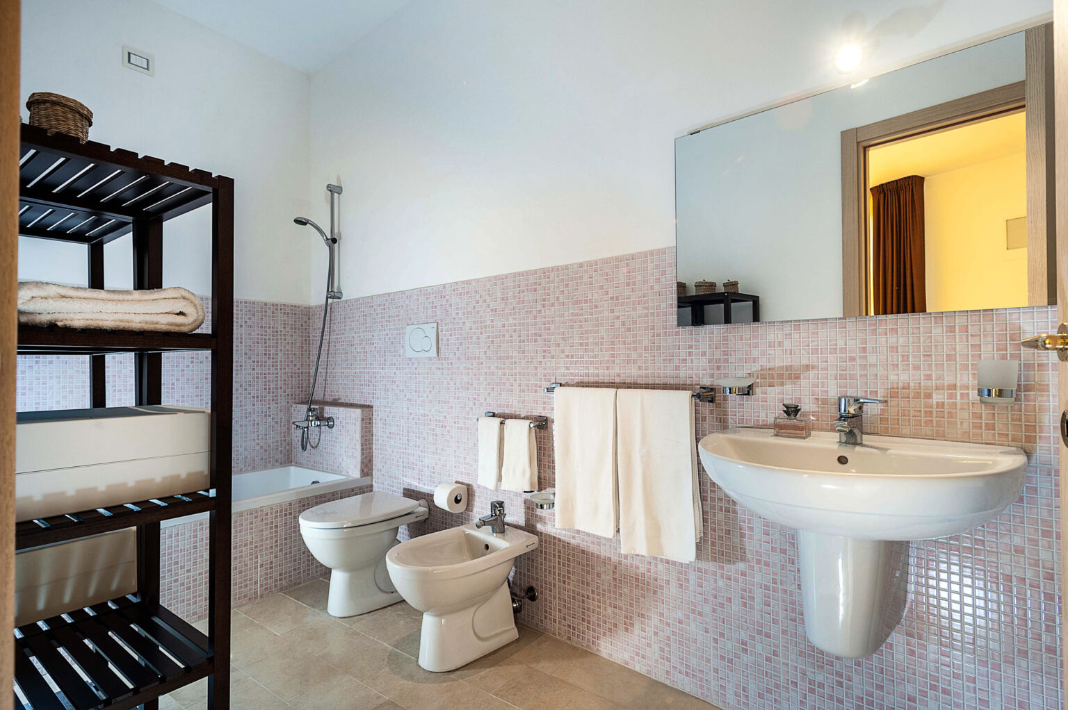Modern bathroom featuring a shower, bathtub, sink, and bidet, complemented by a stylish shelving unit with neatly arranged towels. The walls are adorned with light pink tiles, creating a clean and inviting atmosphere. | Only on SopranoVillas