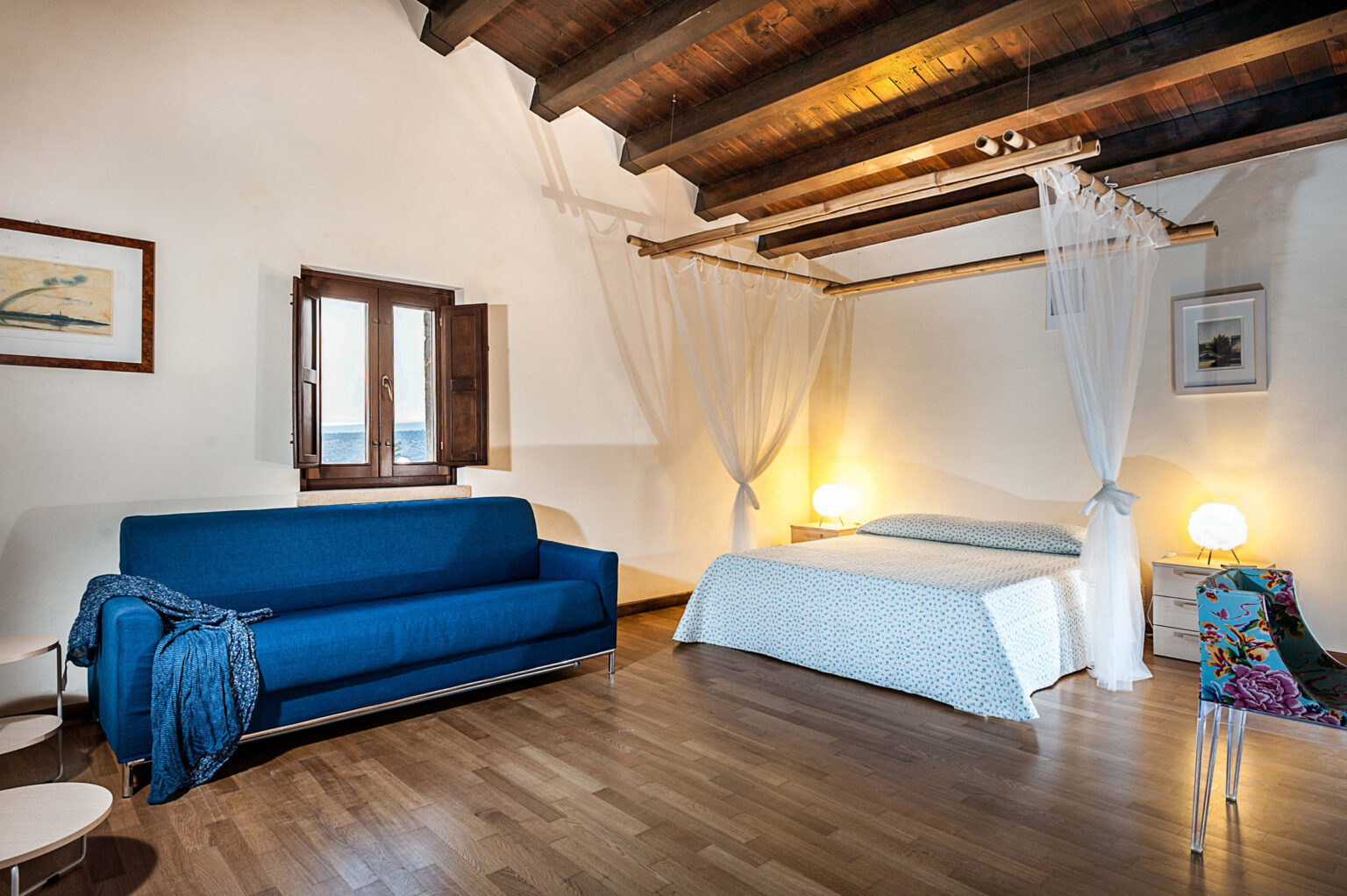Cozy bedroom interior featuring a blue sofa, a double bed with light bedding, wooden ceiling beams, and a window with open shutters. Soft lighting from bedside lamps creates a warm atmosphere, complemented by decorative elements like a patterned chair and artwork on the walls. Ideal for relaxation and comfort. | Only on SopranoVillas