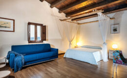 Cozy bedroom interior featuring a blue sofa, a double bed with light bedding, wooden ceiling beams, and a window with open shutters. Soft lighting from bedside lamps creates a warm atmosphere, complemented by decorative elements like a patterned chair and artwork on the walls. Ideal for relaxation and comfort.
