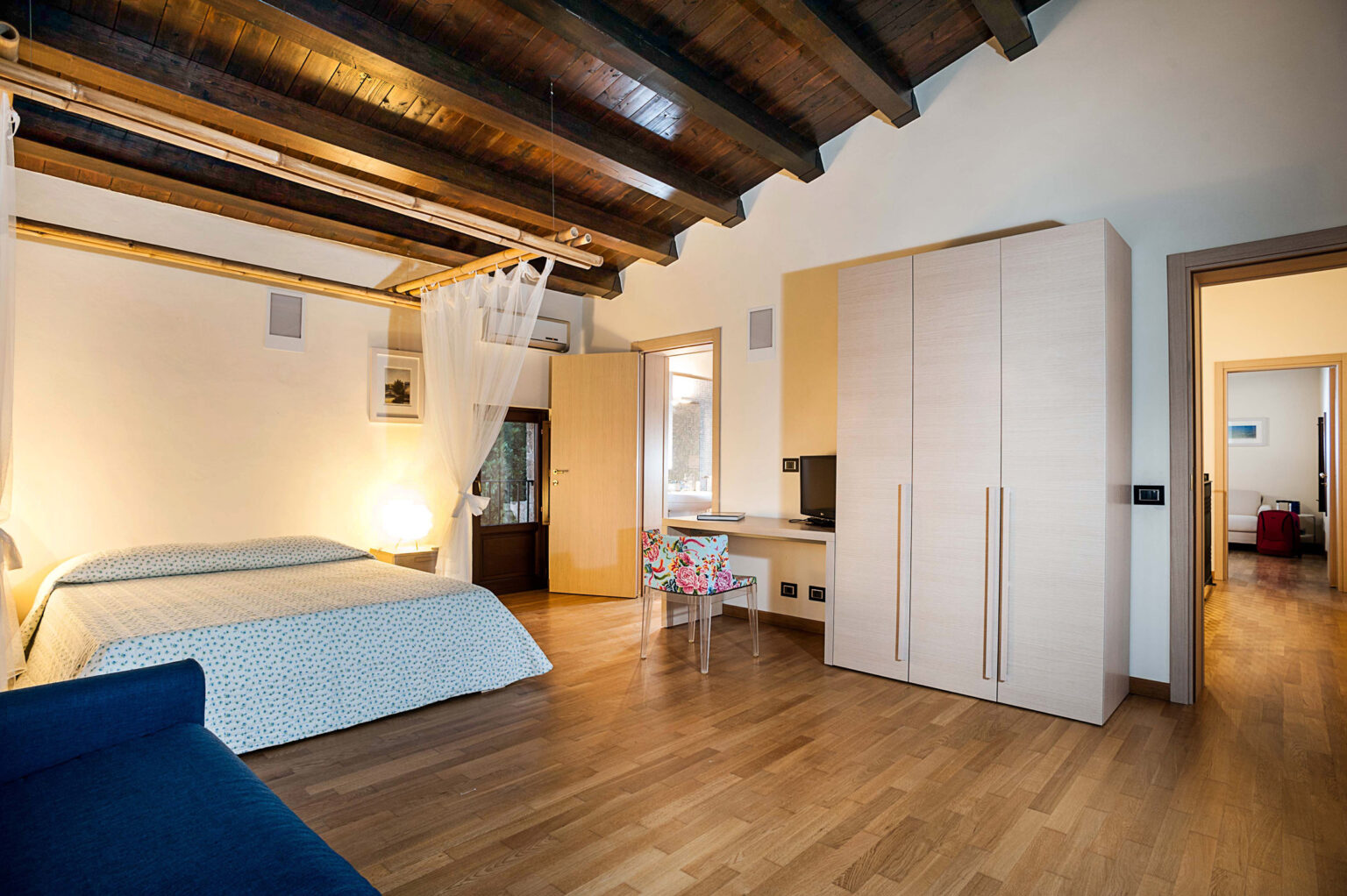 Modern bedroom interior featuring wooden beams, a double bed with light blue bedding, a wardrobe, and a small desk. The room is well-lit with natural light and has a cozy, inviting atmosphere. | Only on SopranoVillas