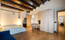 Modern bedroom interior featuring wooden beams, a double bed with light blue bedding, a wardrobe, and a small desk. The room is well-lit with natural light and has a cozy, inviting atmosphere.