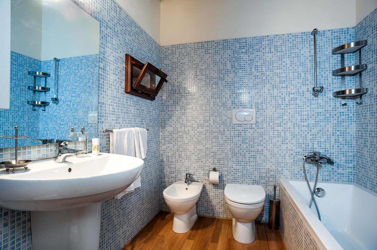 Modern bathroom featuring blue mosaic tiles, a white sink, bidet, and toilet, along with a bathtub. The space includes a wooden mirror and towel rack, highlighting a clean and contemporary design. | Only on SopranoVillas