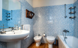 Modern bathroom featuring blue mosaic tiles, a white sink, bidet, and toilet, along with a bathtub. The space includes a wooden mirror and towel rack, highlighting a clean and contemporary design.