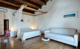 Cozy bedroom interior featuring a double bed with a canopy, a comfortable sofa, and warm wooden beams, complemented by soft lighting and minimalist decor.