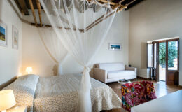 Cozy bedroom featuring a king-sized bed with a white canopy, a comfortable sofa, and large windows that allow natural light. Decor includes colorful accents and framed artwork, creating a warm and inviting atmosphere.