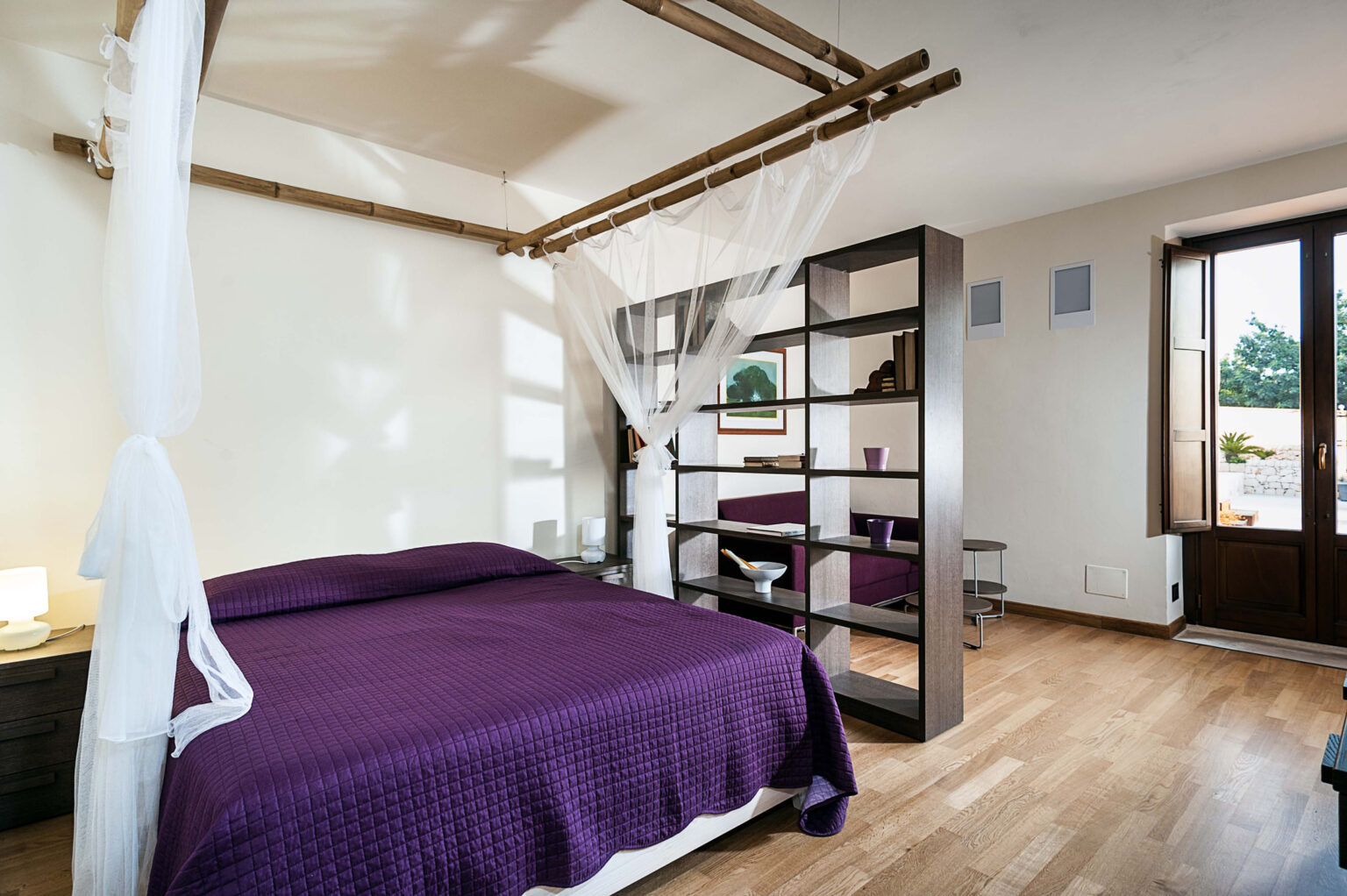 Modern bedroom featuring a purple bedspread, bamboo canopy, wooden flooring, and a stylish bookshelf. Natural light streams in through large windows, creating a bright and inviting atmosphere. | Only on SopranoVillas