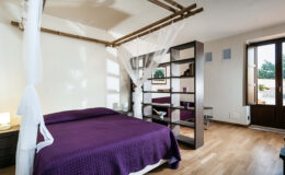 Modern bedroom featuring a purple bedspread, bamboo canopy, wooden flooring, and a stylish bookshelf. Natural light streams in through large windows, creating a bright and inviting atmosphere.
