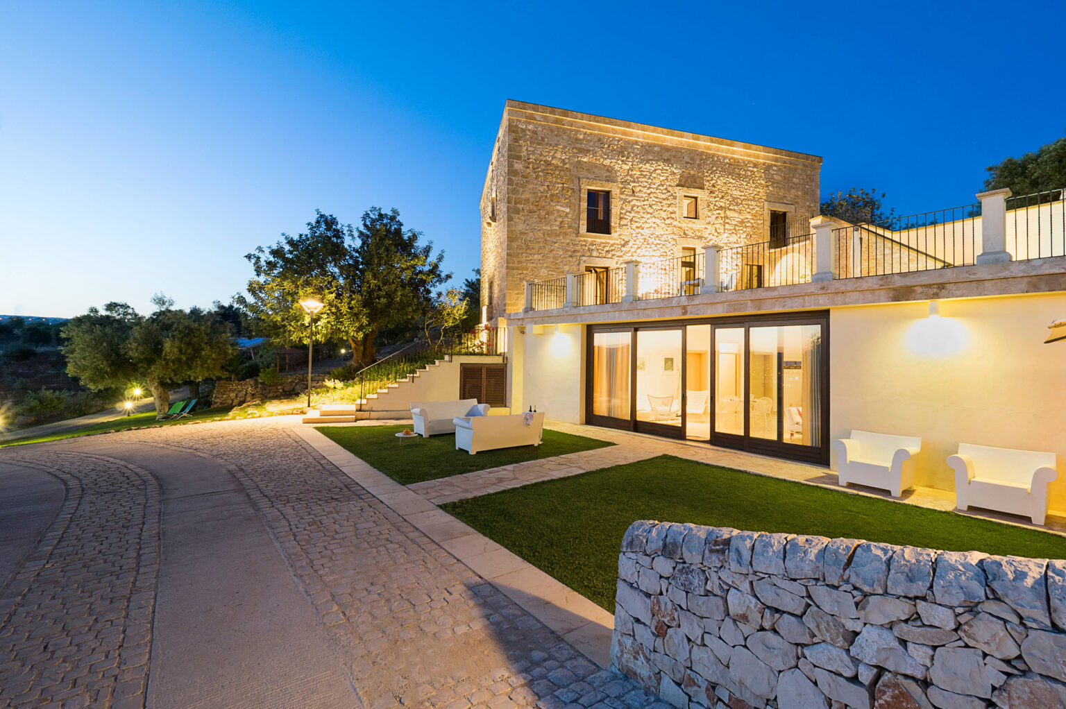 Modern stone villa illuminated at dusk, featuring a landscaped garden, outdoor seating, and a cobblestone driveway, set against a clear blue sky. | Only on SopranoVillas