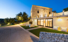 Modern stone villa illuminated at dusk, featuring a landscaped garden, outdoor seating, and a cobblestone driveway, set against a clear blue sky.