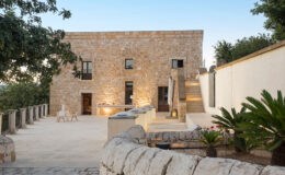 Exterior view of a modern stone villa with a spacious patio, featuring elegant outdoor seating and lush landscaping, highlighting a blend of contemporary and rustic architectural styles.