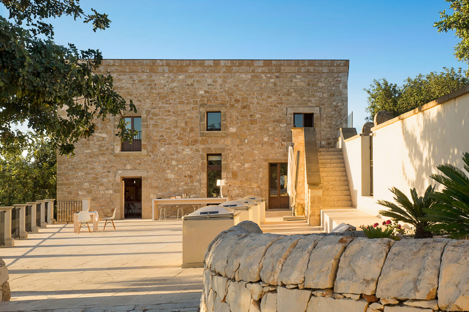 Stone house exterior with a spacious terrace, featuring modern outdoor furniture, stairs leading to an upper level, and lush greenery surrounding the property. The image showcases a blend of traditional architecture and contemporary design elements. | Only on SopranoVillas