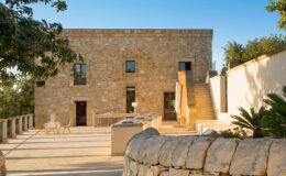 Stone house exterior with a spacious terrace, featuring modern outdoor furniture, stairs leading to an upper level, and lush greenery surrounding the property. The image showcases a blend of traditional architecture and contemporary design elements.
