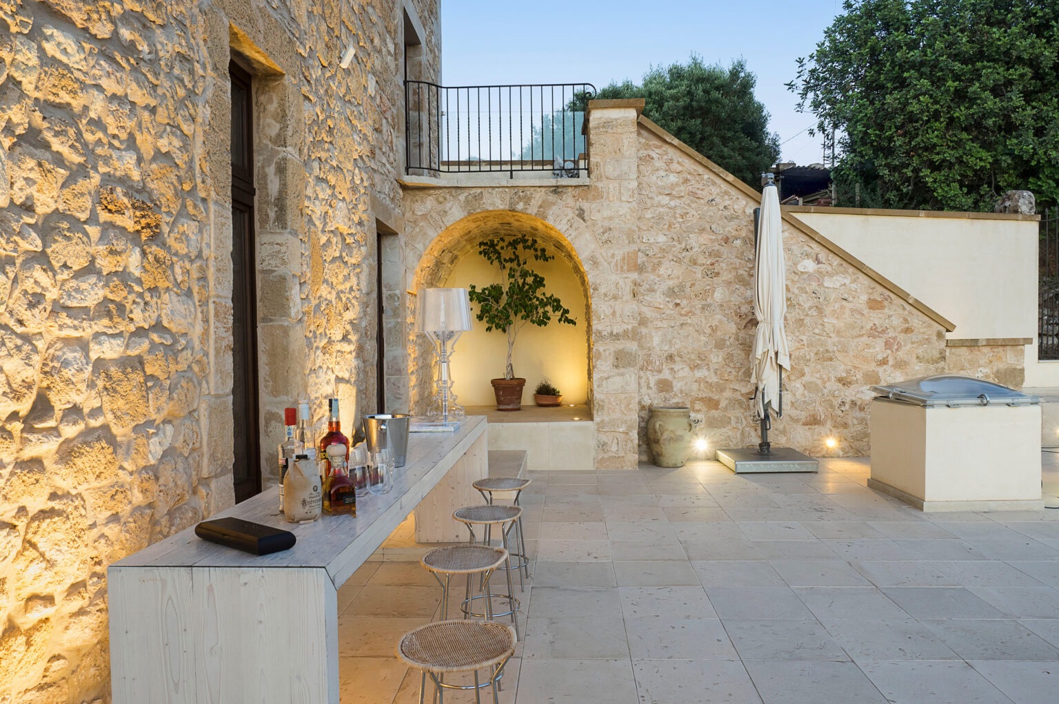Outdoor patio area featuring a stone wall, a modern bar with various bottles and glasses, and stylish seating. A potted plant adds greenery, while warm lighting creates an inviting atmosphere. Ideal for entertaining and relaxation in a serene environment. | Only on SopranoVillas