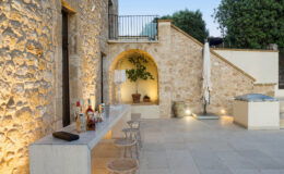 Outdoor patio area featuring a stone wall, a modern bar with various bottles and glasses, and stylish seating. A potted plant adds greenery, while warm lighting creates an inviting atmosphere. Ideal for entertaining and relaxation in a serene environment.