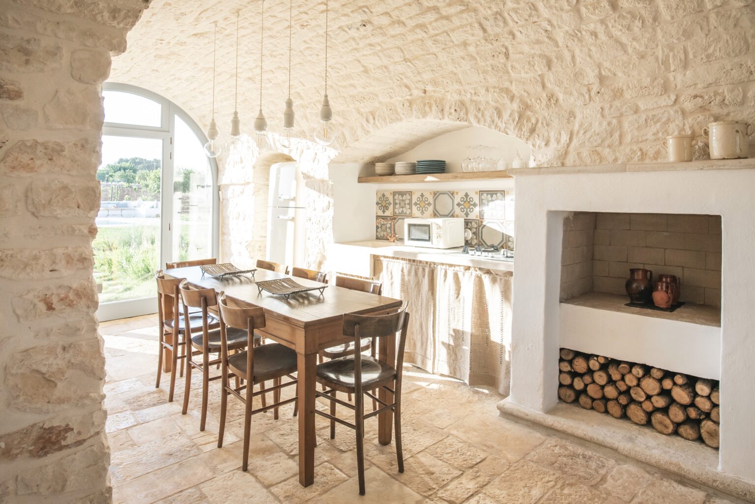 Bright and airy kitchen with stone walls, wooden dining table, and modern appliances, featuring a cozy fireplace and stacked firewood. Natural light floods the space through large windows, creating a warm and inviting atmosphere. | Only on SopranoVillas