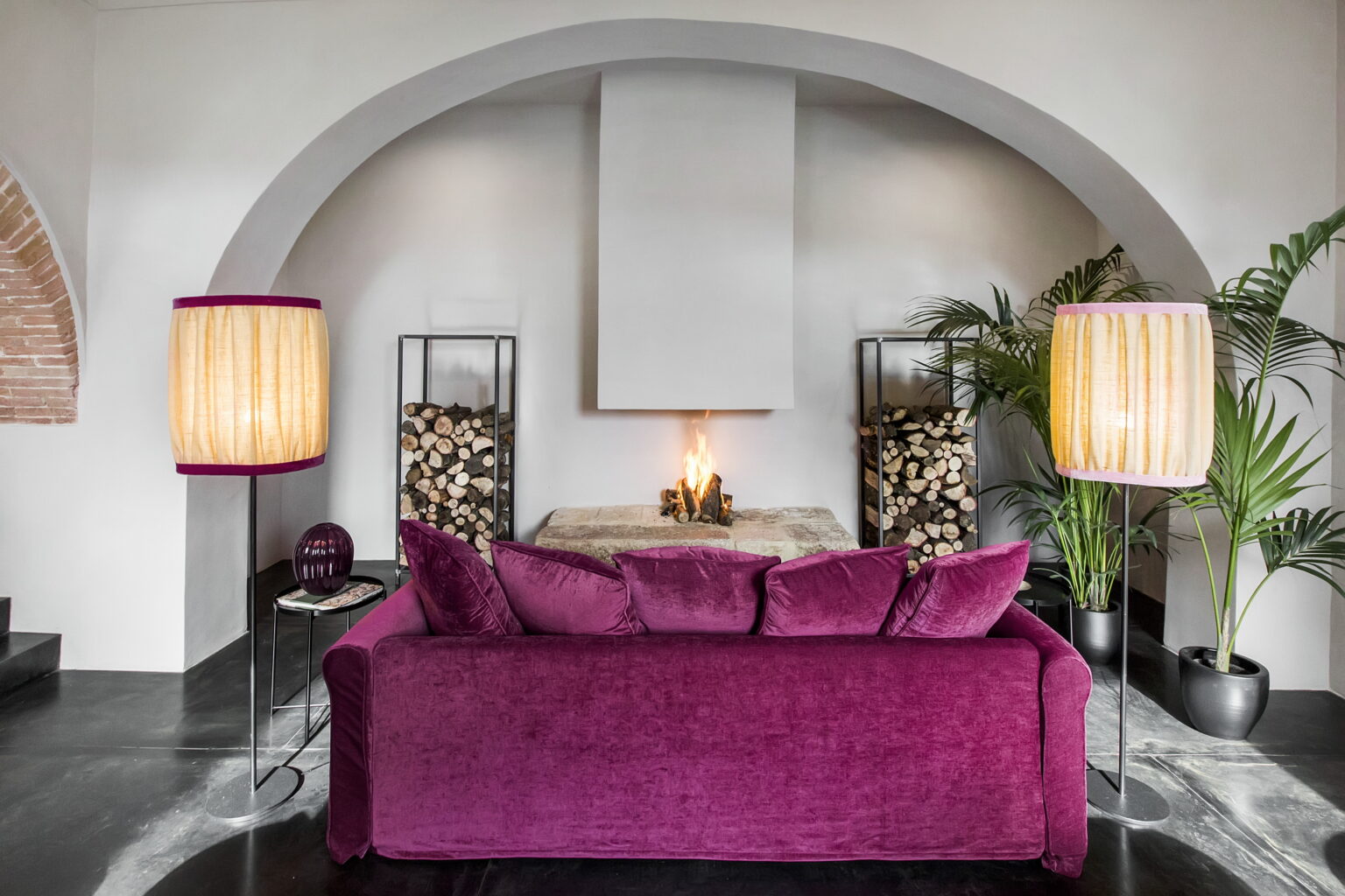 Modern living room featuring a plush purple sofa, stylish floor lamps, and decorative plants, creating a cozy and inviting atmosphere. The design includes an elegant archway and a minimalist fireplace, enhancing the contemporary aesthetic. | Only on SopranoVillas