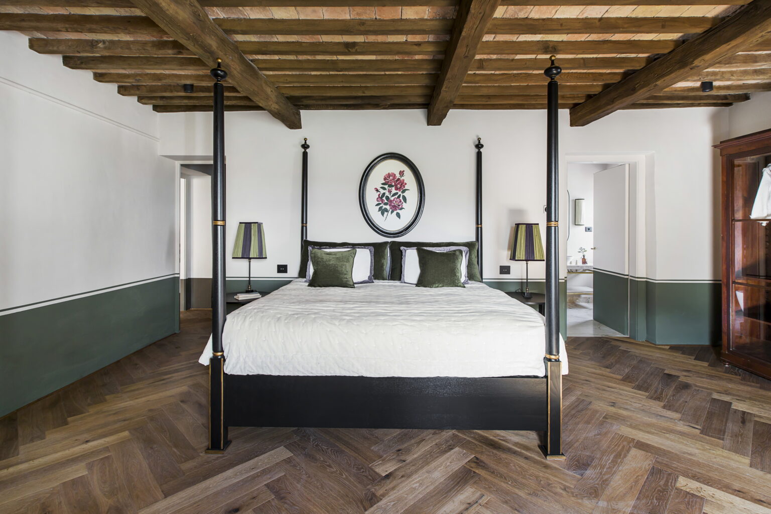 Elegant bedroom featuring a four-poster bed with a white bedding set, decorative pillows, and stylish lamps. The room showcases rustic wooden beams and herringbone wood flooring, complemented by a green accent wall and a framed artwork above the bed, creating a cozy and sophisticated atmosphere. | Only on SopranoVillas