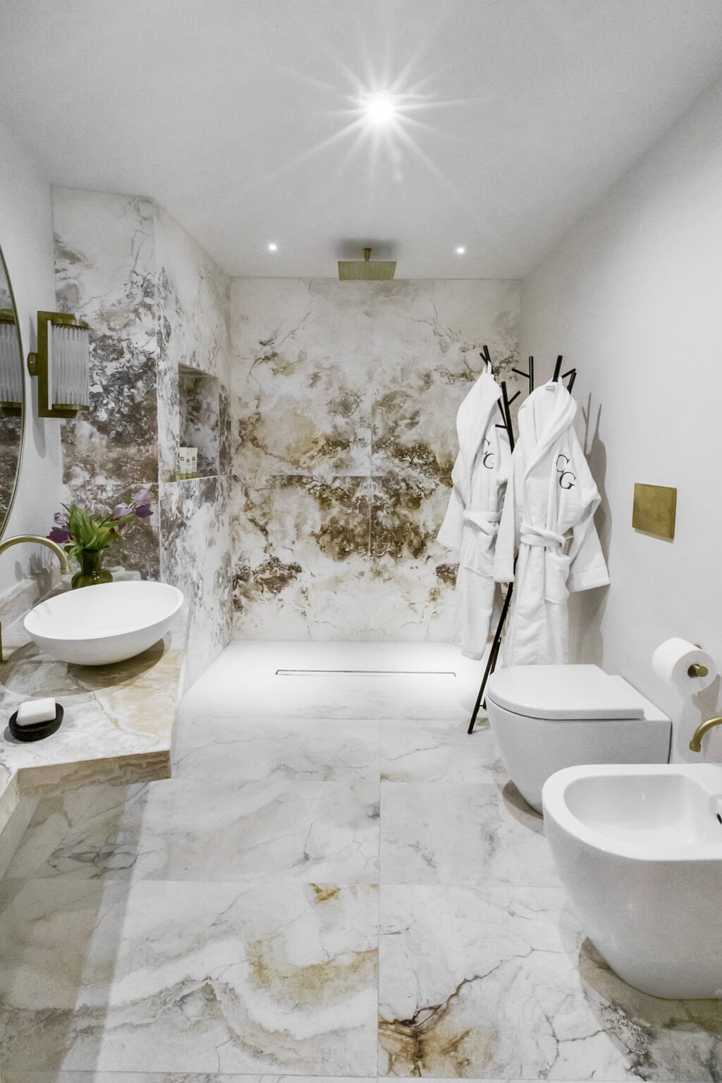 Modern bathroom featuring a stylish design with marble walls, a sleek toilet, a washbasin, and plush white bathrobes hanging on a rack, creating a luxurious and inviting atmosphere. | Only on SopranoVillas