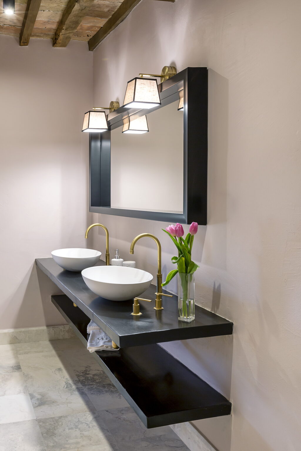 Modern bathroom design featuring two white vessel sinks, elegant gold faucets, and a stylish mirror above a dark countertop. A vase with pink tulips adds a touch of color to the space. | Only on SopranoVillas