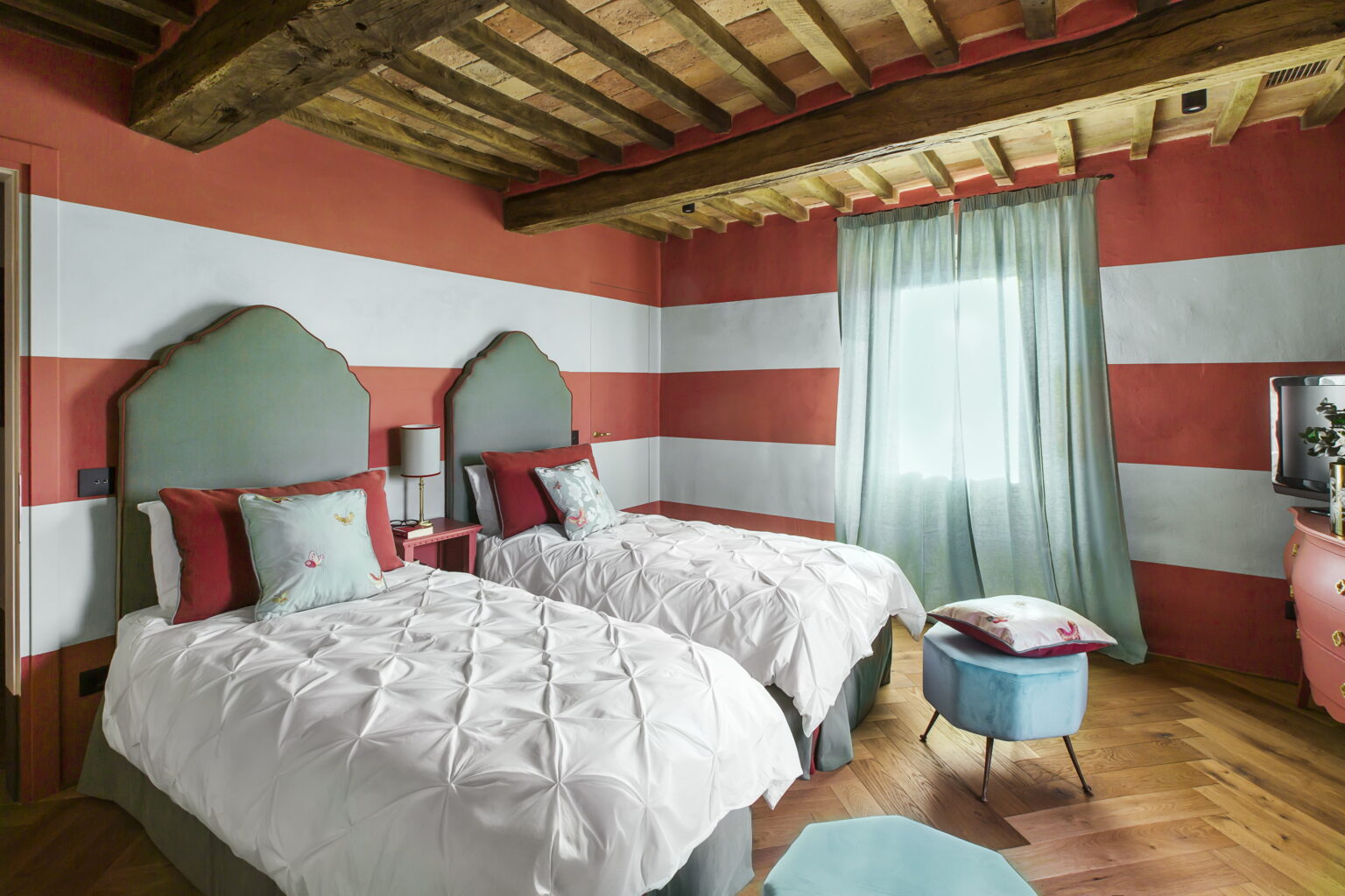 Cozy guest bedroom featuring two twin beds with white bedding, accented by red and teal pillows. The walls are painted in a warm coral hue with white stripes, and natural light filters through a sheer curtain, creating a welcoming atmosphere. A stylish blue stool adds a pop of color to the wooden floor. | Only on SopranoVillas
