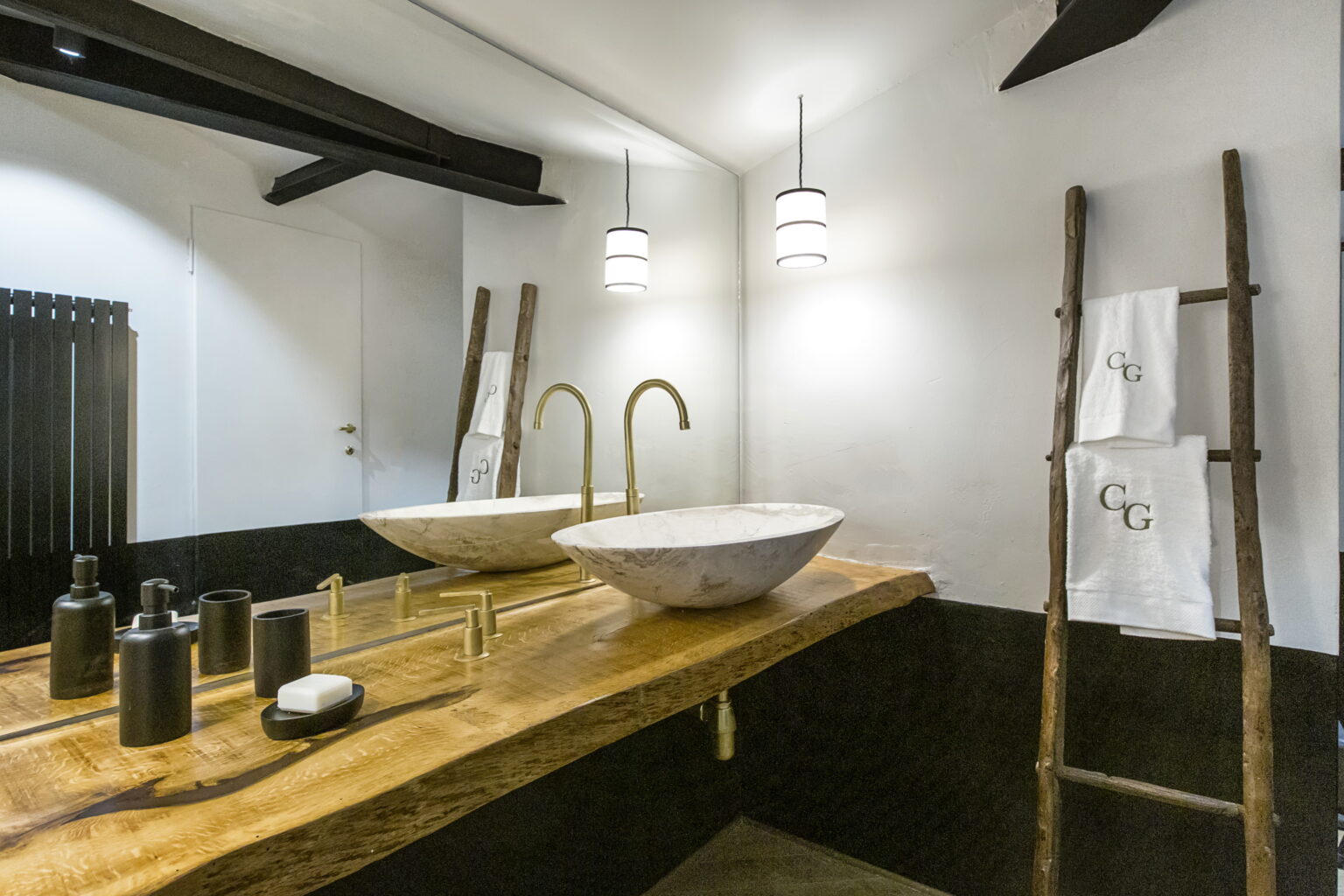 Modern bathroom interior featuring a wooden countertop with two stone sinks, stylish black accessories, and hanging pendant lights. A wooden ladder displays neatly folded towels, enhancing the contemporary aesthetic. | Only on SopranoVillas