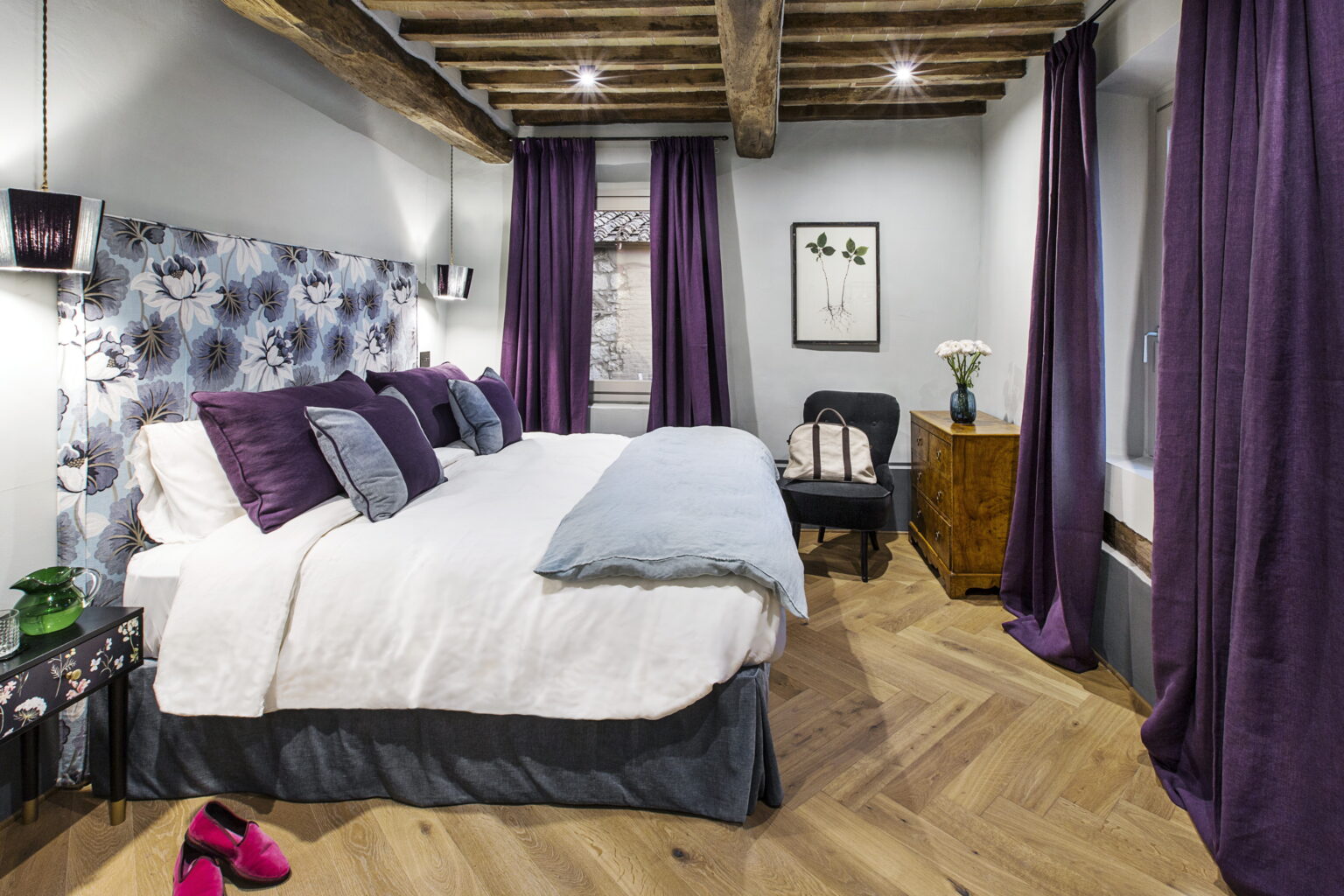 Cozy bedroom featuring a king-size bed with plush purple pillows, elegant wood beams, and stylish decor. The room includes a comfortable chair, a wooden nightstand, and rich purple curtains, creating a warm and inviting atmosphere. | Only on SopranoVillas