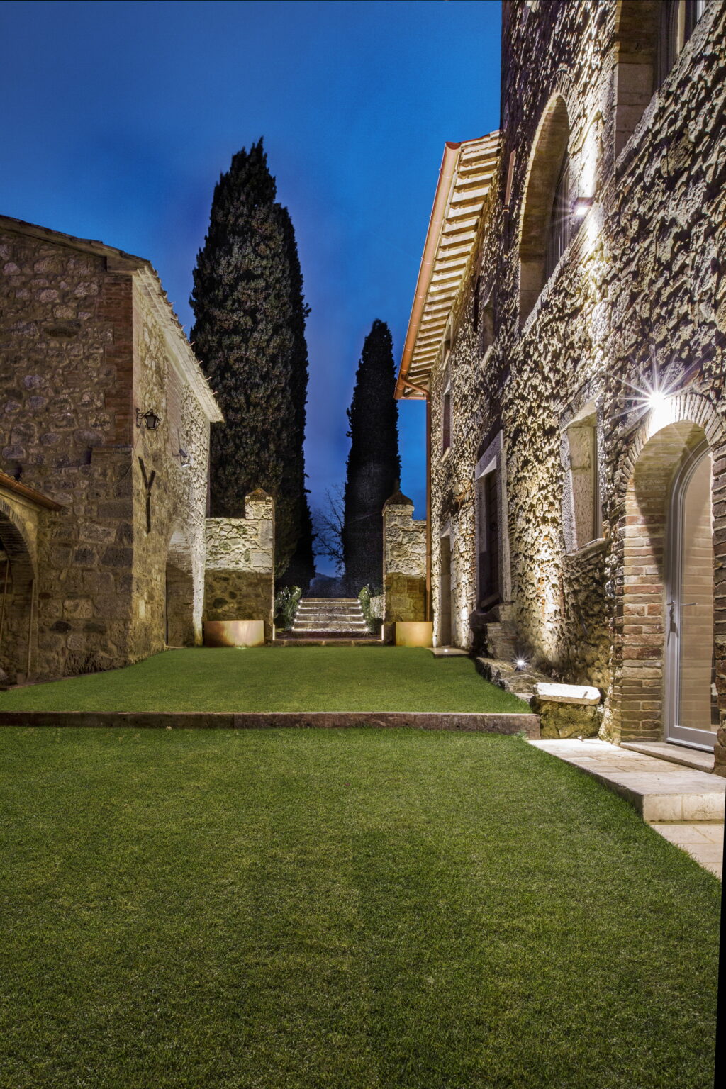 Stone courtyard at dusk with illuminated pathways and lush green grass, framed by tall cypress trees and rustic stone buildings, creating a serene outdoor ambiance. | Only on SopranoVillas