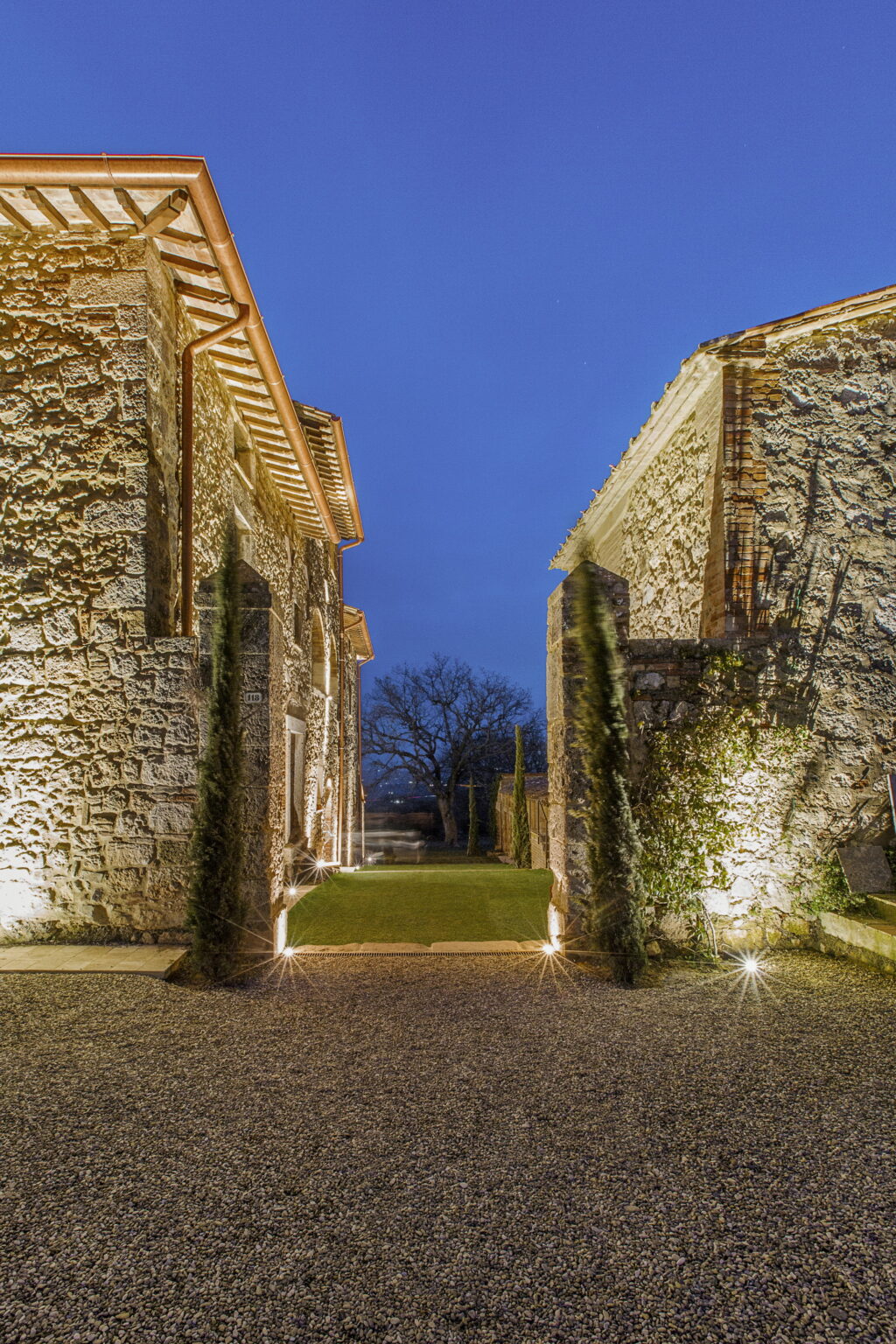 Stone pathway between illuminated rustic buildings at dusk, surrounded by greenery and a tree in the background. | Only on SopranoVillas