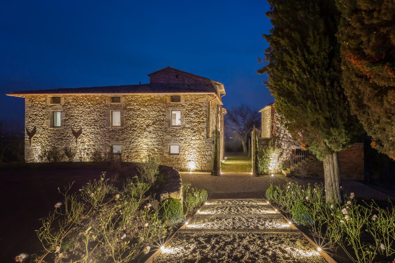 Stone villa illuminated at night, featuring a landscaped pathway leading to the entrance, surrounded by greenery and cypress trees. Perfect for travelers seeking a serene getaway in a rustic setting. | Only on SopranoVillas