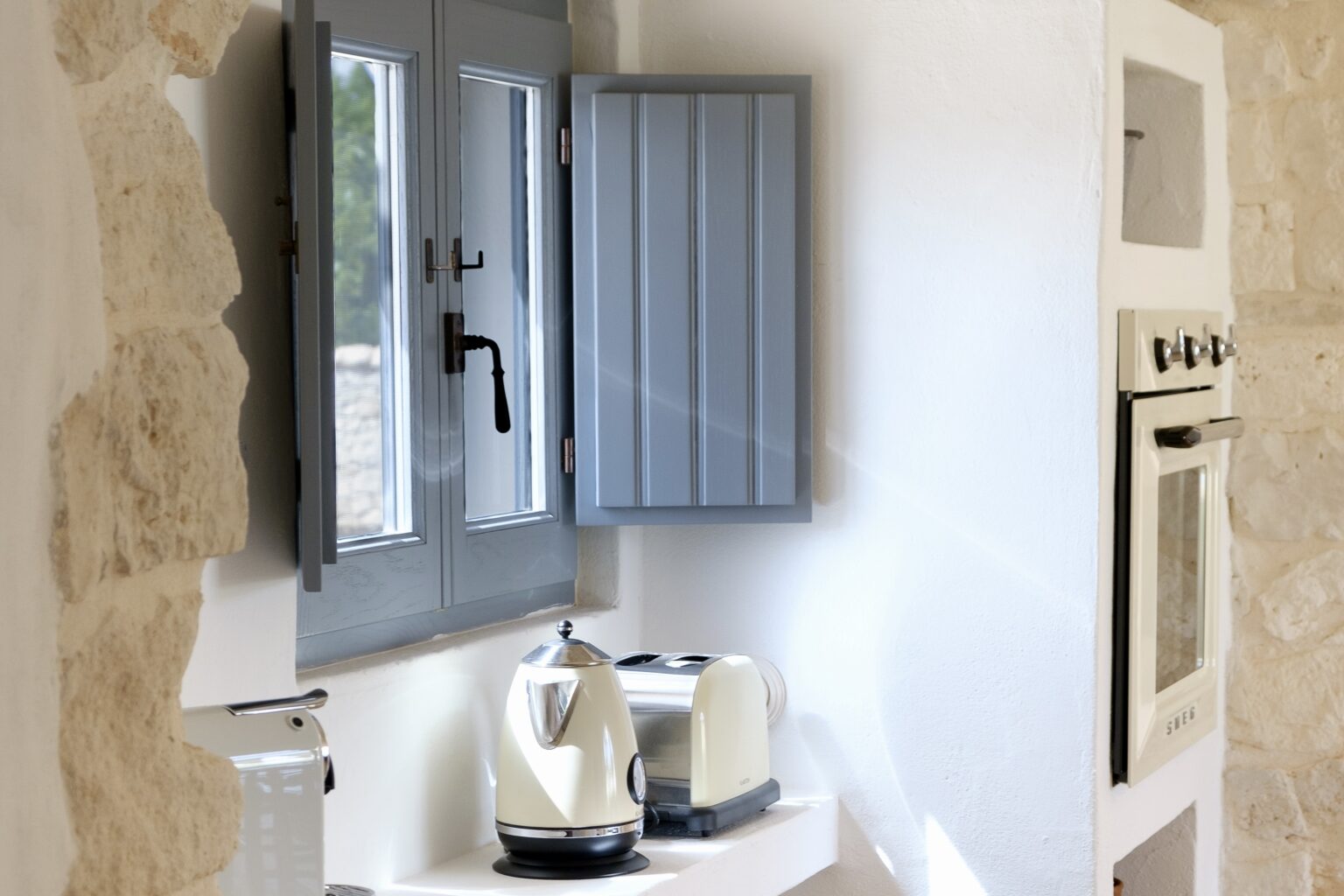 Modern kitchen interior featuring a gray shuttered window, stainless steel kettle, and a white countertop, complemented by a stone wall and built-in oven. | Only on SopranoVillas