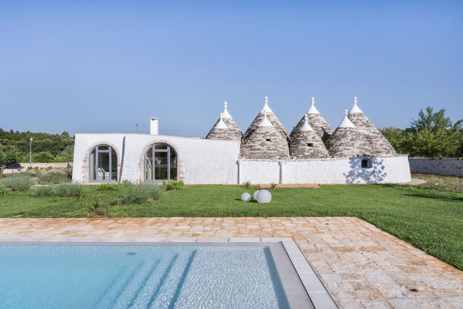 Trullo-style house with conical roofs surrounded by green lawn and a swimming pool under a clear blue sky. | Only on SopranoVillas