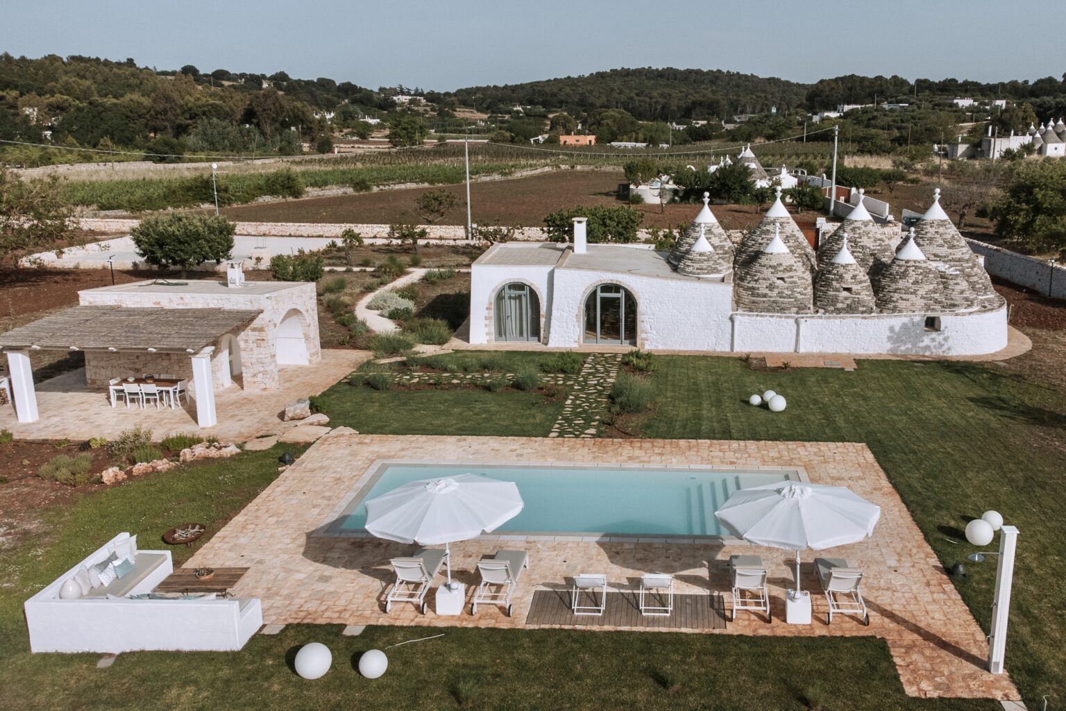 Aerial view of a luxurious villa featuring traditional trulli architecture, surrounded by lush greenery and farmland. The property includes a sparkling swimming pool with sun loungers and umbrellas, highlighting a serene outdoor living space perfect for relaxation and entertainment. | Only on SopranoVillas