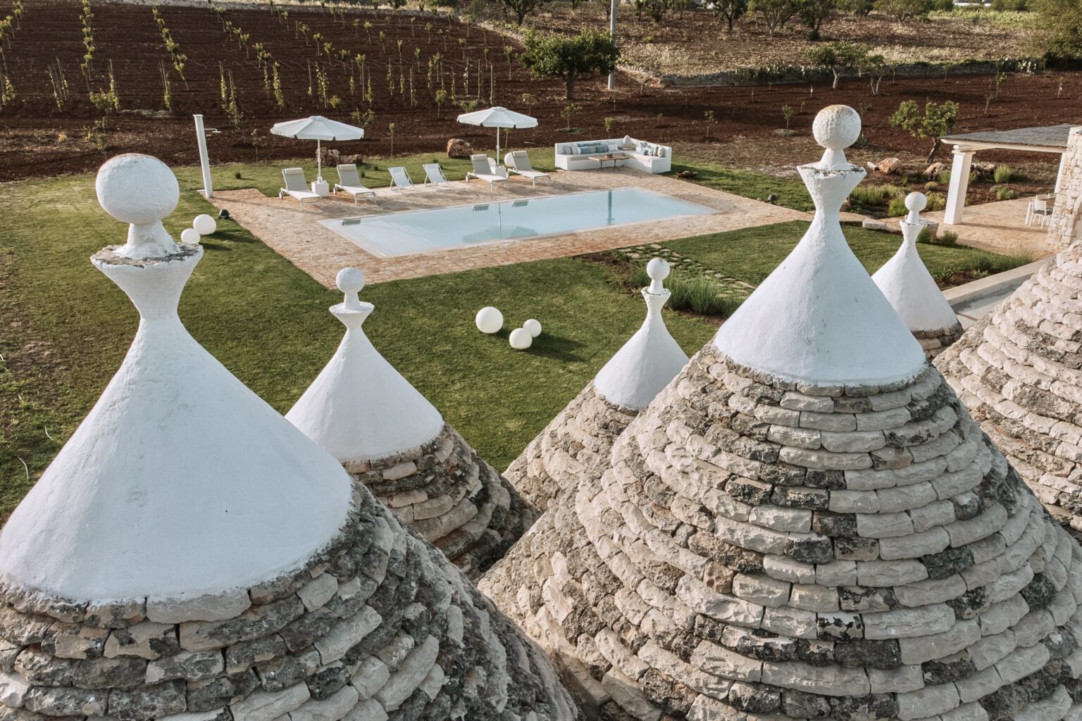 Trulli houses with conical roofs in the foreground, overlooking a luxurious pool area with lounge chairs and umbrellas, set against a backdrop of terraced vineyards. | Only on SopranoVillas