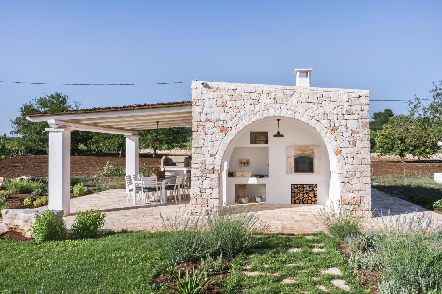 Charming outdoor stone kitchen and dining area featuring a white stone structure with an arched entrance, surrounded by lush greenery and a clear blue sky. Ideal for entertaining and enjoying alfresco meals. | Only on SopranoVillas