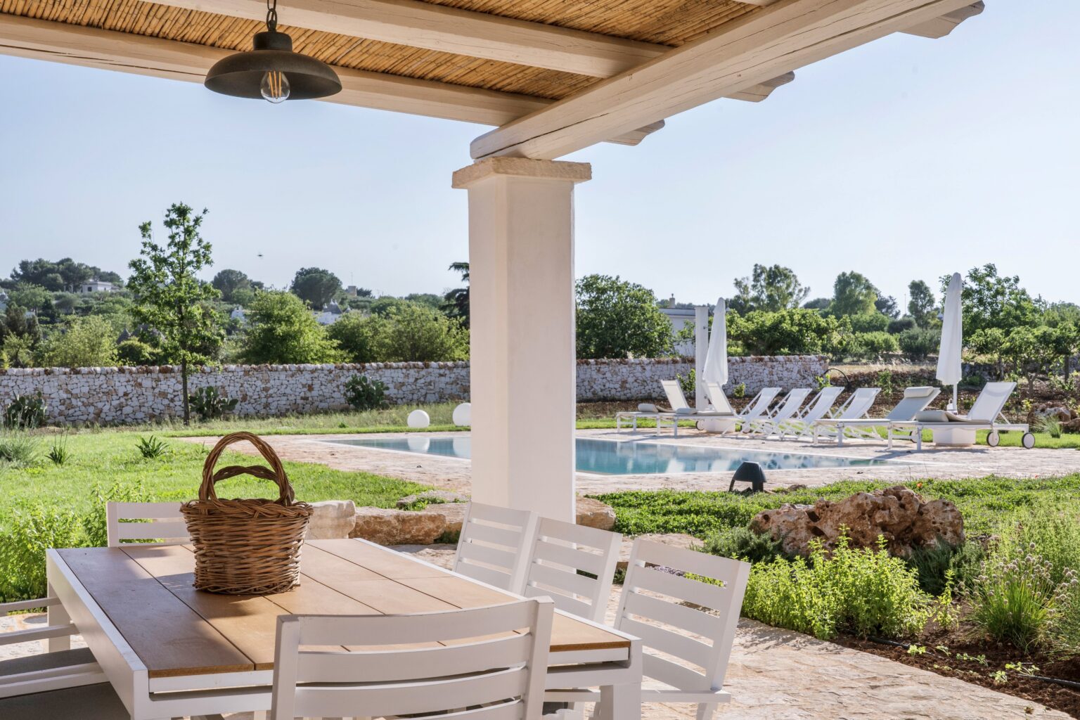 Outdoor dining area with a wooden table and basket, overlooking a swimming pool surrounded by lounge chairs and lush greenery in a serene setting. | Only on SopranoVillas