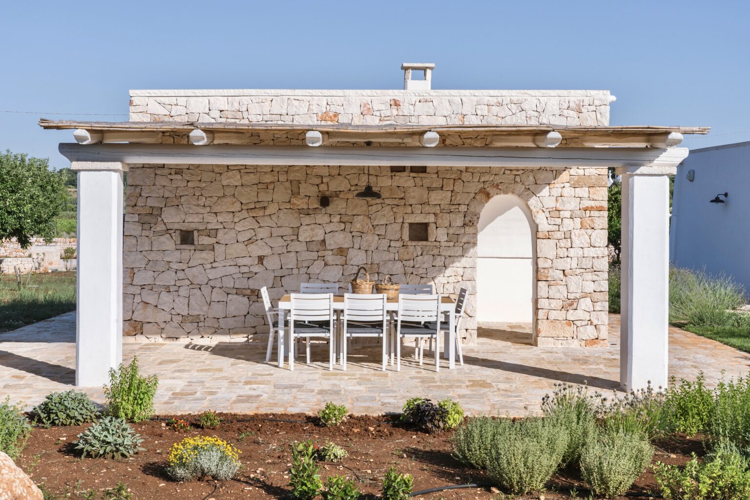 Charming stone cottage with a covered patio and dining table, surrounded by lush greenery and flowers, set against a clear blue sky. Ideal for outdoor gatherings and relaxation. | Only on SopranoVillas