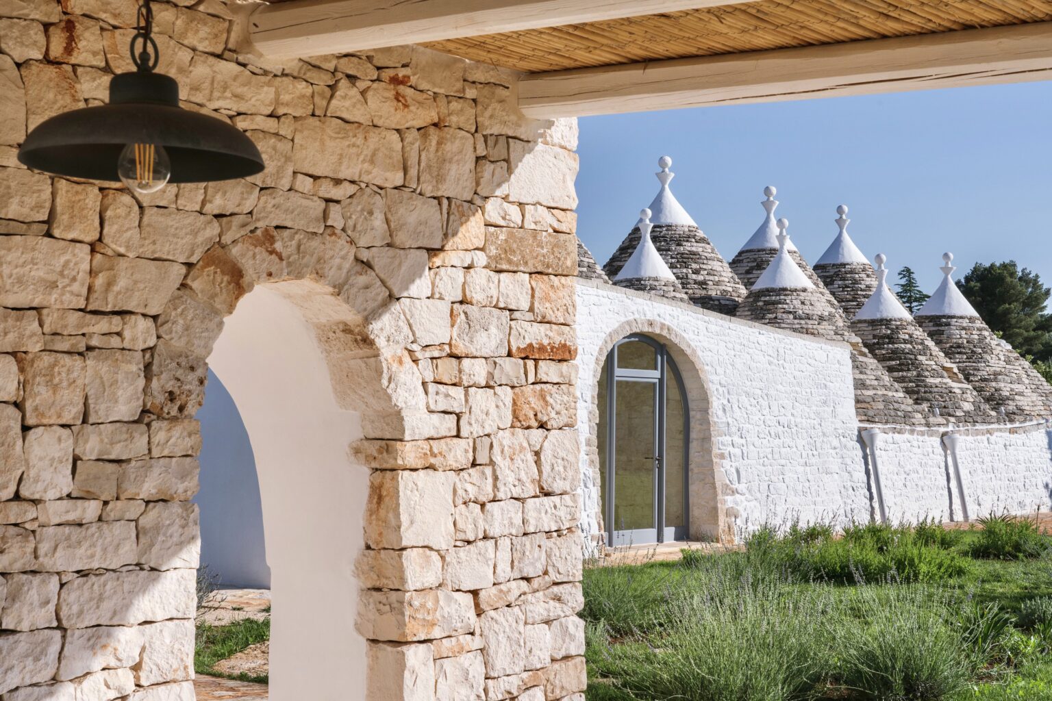 Trullo houses with conical roofs in Puglia, Italy, framed by an archway and surrounded by lush greenery under a clear blue sky. | Only on SopranoVillas