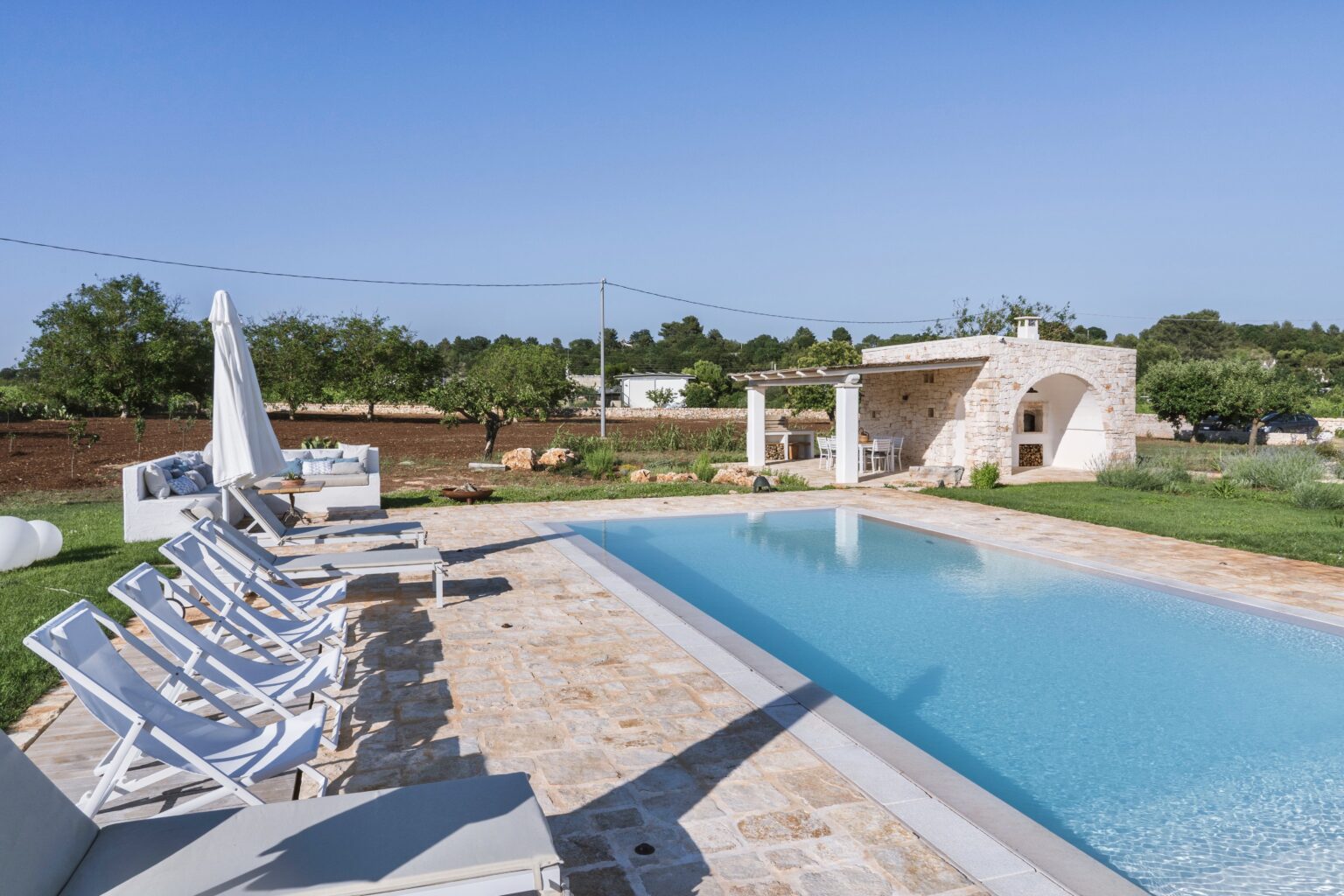 Luxury outdoor swimming pool surrounded by sun loungers in a serene landscape with olive trees and a stone structure in the background. Ideal for relaxation and leisure activities. | Only on SopranoVillas