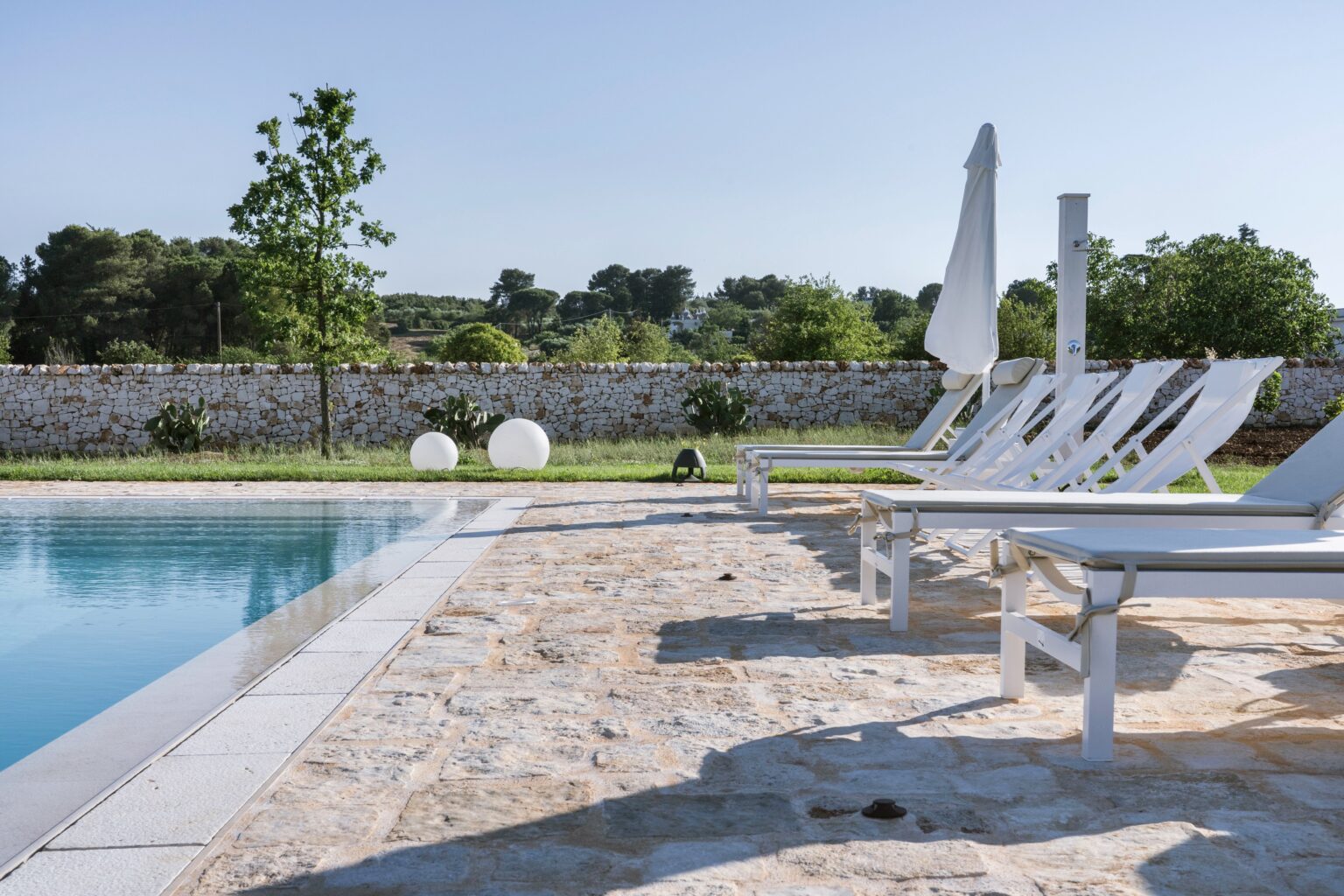 Modern outdoor swimming pool area with white lounge chairs, surrounded by lush greenery and a stone wall, ideal for relaxation and leisure. | Only on SopranoVillas