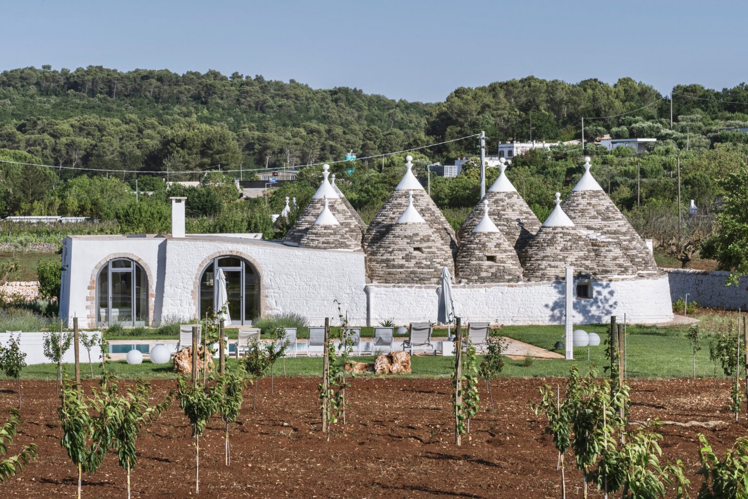 Trulli-style houses surrounded by lush greenery and vineyards, featuring a modern white building with large windows and a pool area, set against a backdrop of rolling hills in a picturesque landscape. | Only on SopranoVillas