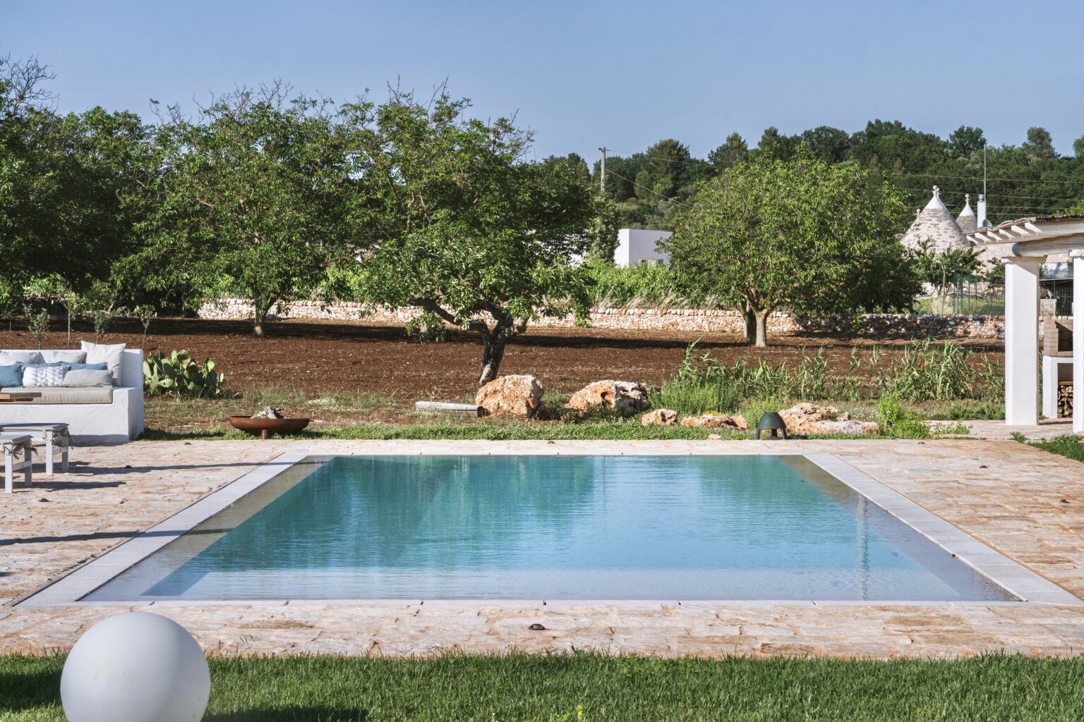 Modern outdoor swimming pool surrounded by lush greenery and stone landscaping, featuring a serene and inviting atmosphere in a sunny setting. | Only on SopranoVillas