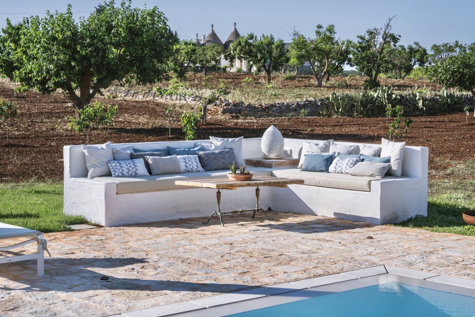 Modern outdoor lounge area featuring a white sectional sofa with cushions, a wooden coffee table, and a scenic backdrop of green trees and farmland, beside a swimming pool. Ideal for relaxing and entertaining in a stylish garden setting. | Only on SopranoVillas